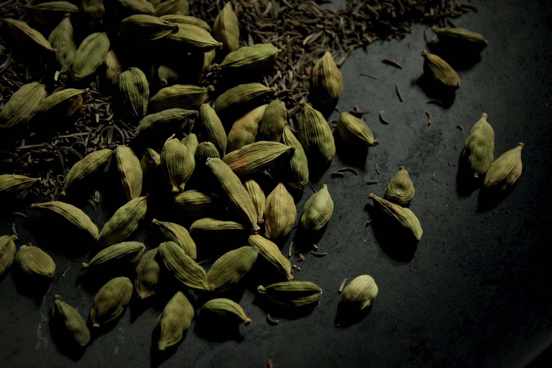 Green cardamom pods and cumin seeds on a dark surface