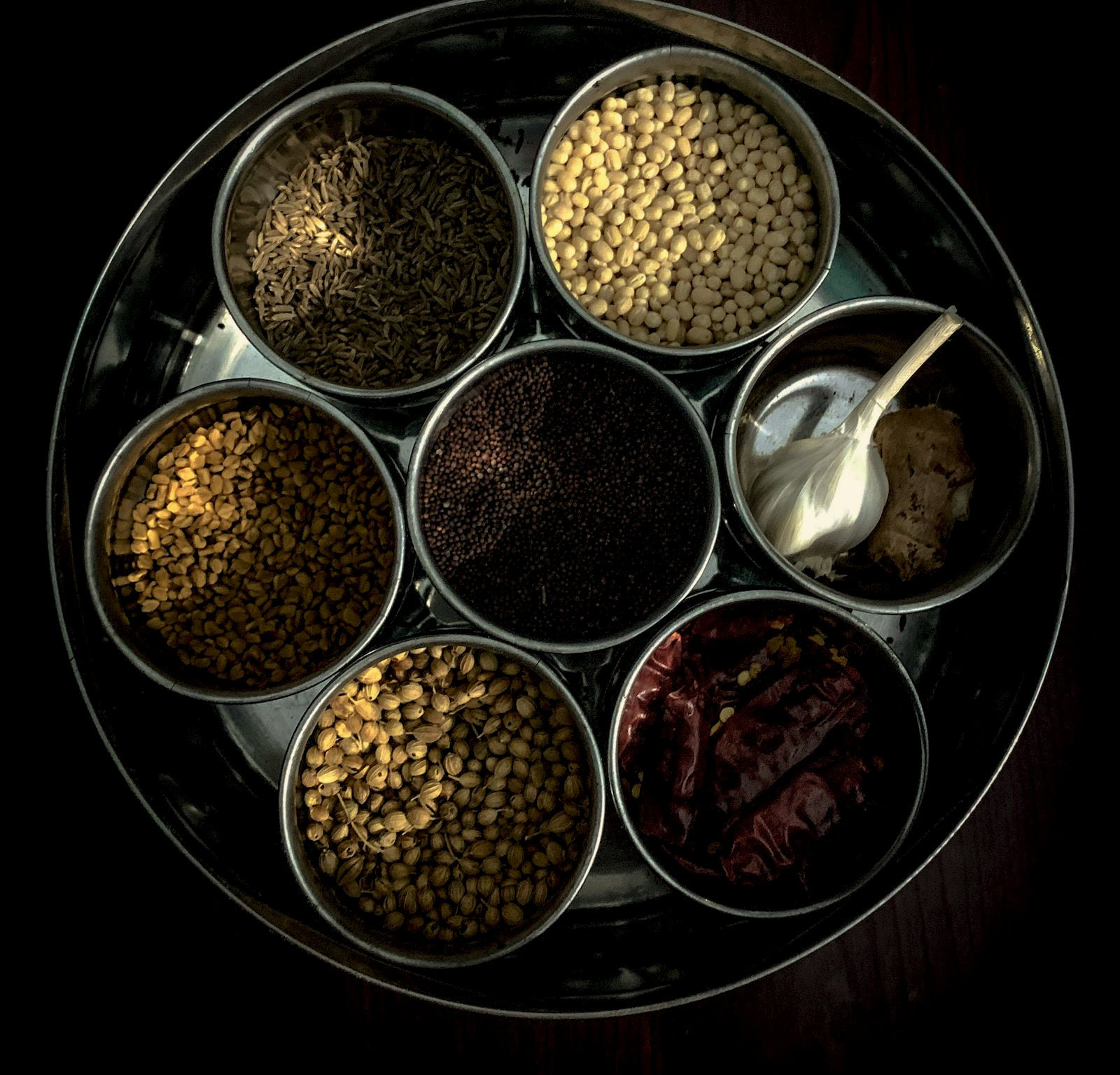 A steel spice box with seven measured compartments of whole spices, lit from above