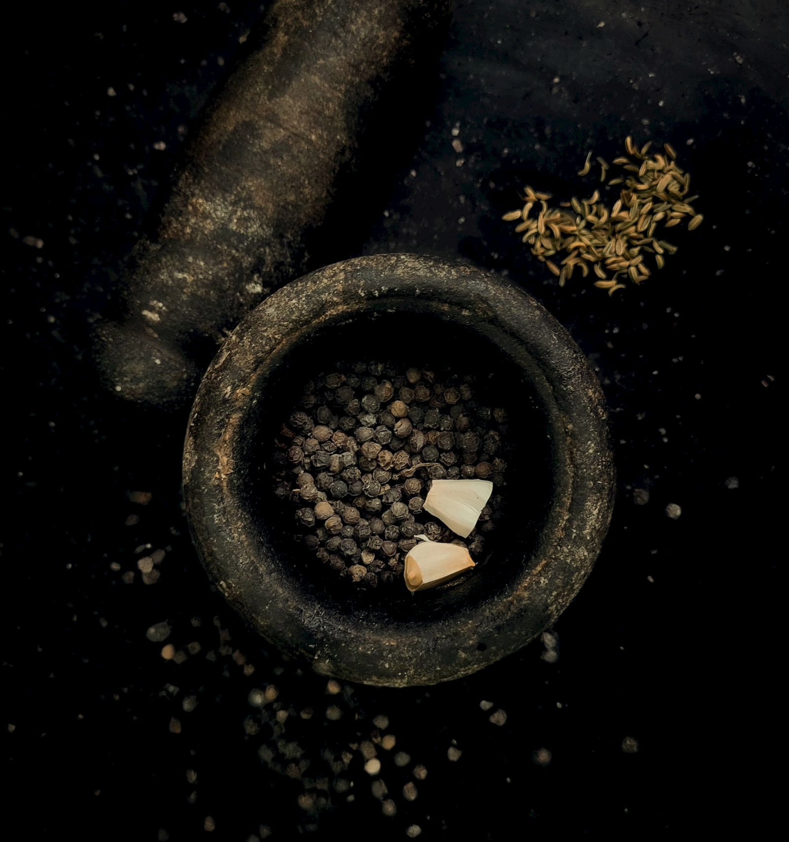 A dark stone mortar holding black peppercorns, with fennel seeds scattered beside it
