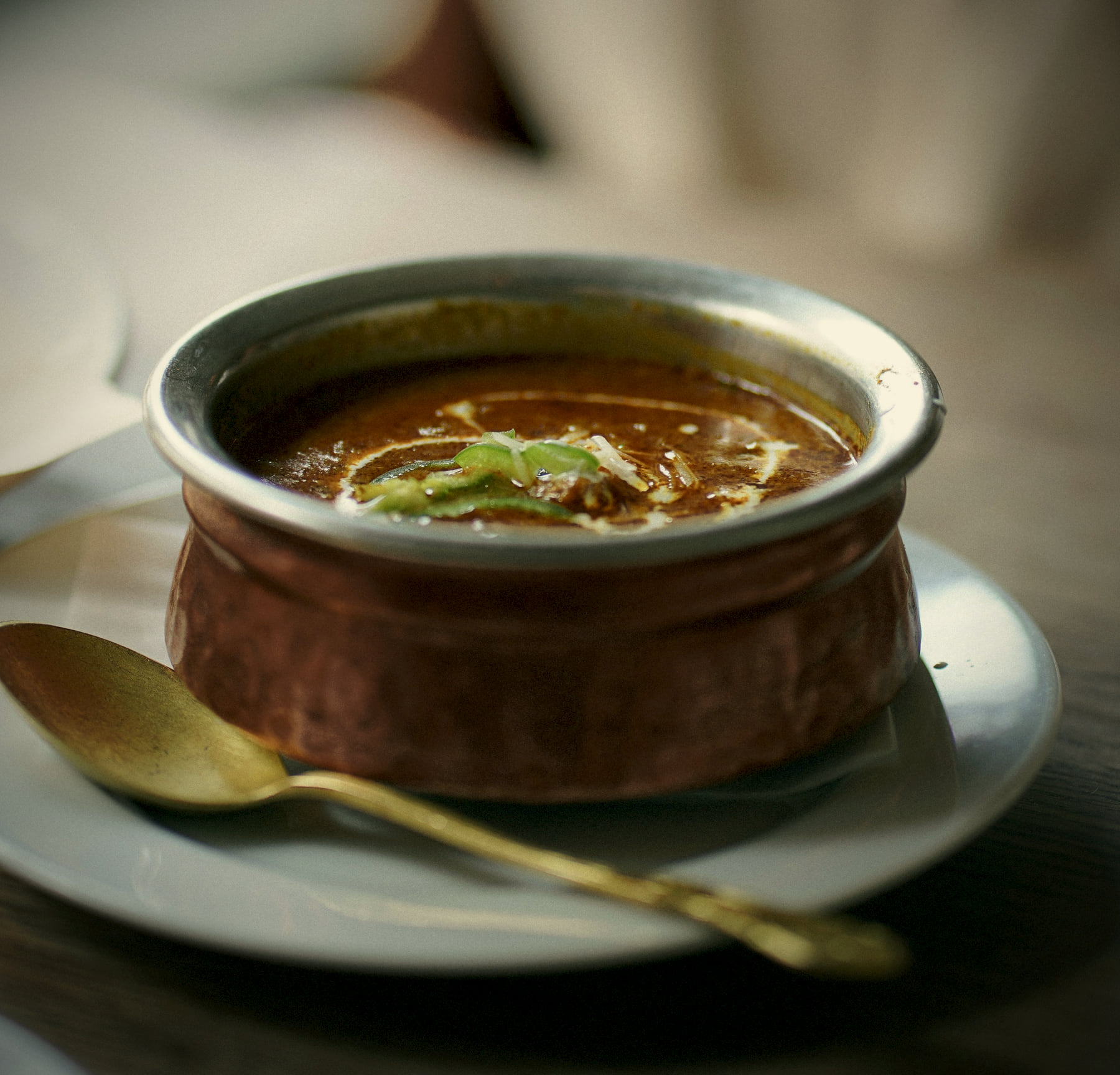 Glossy black dal in a hammered copper handi on an ivory saucer, with a brass spoon beside it