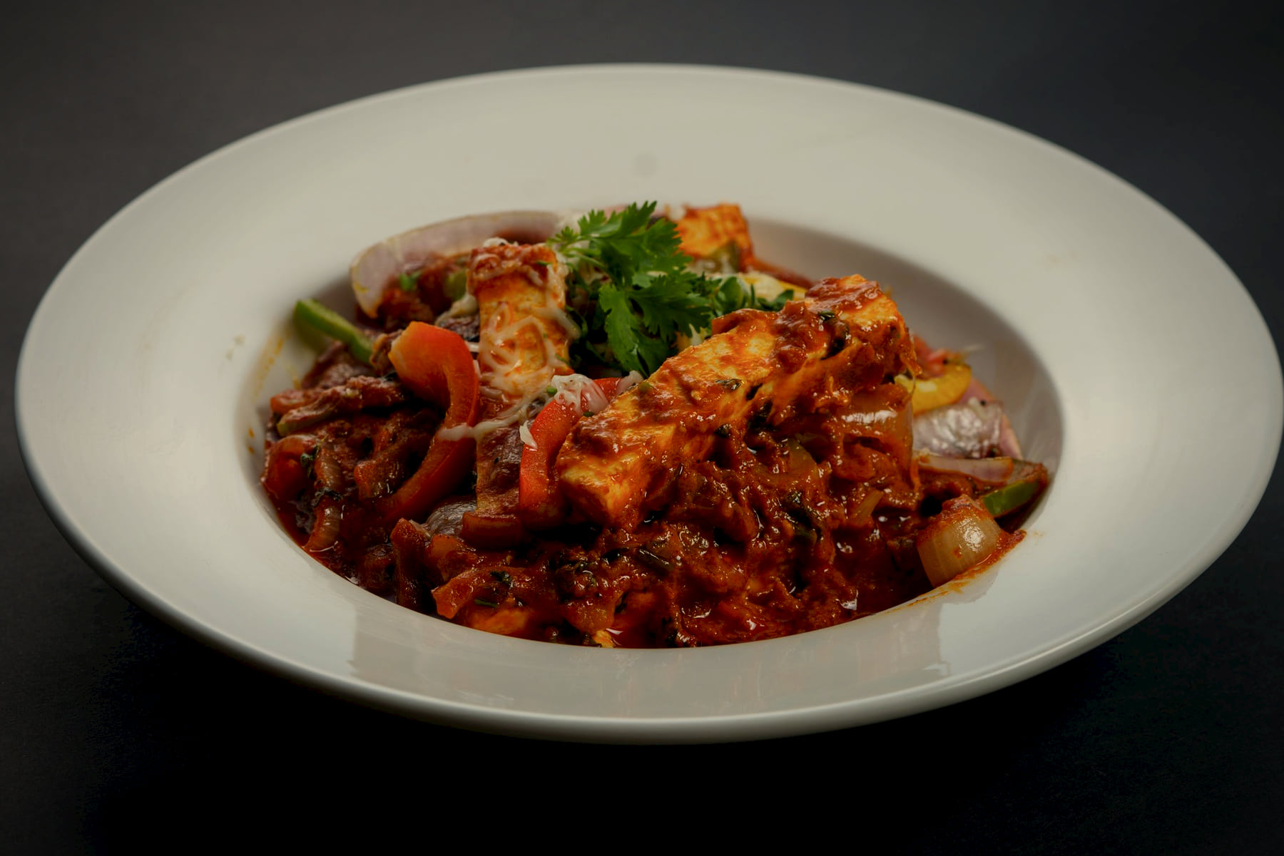 Paneer with onions and peppers in a dark, textured masala, in a wide ivory bowl