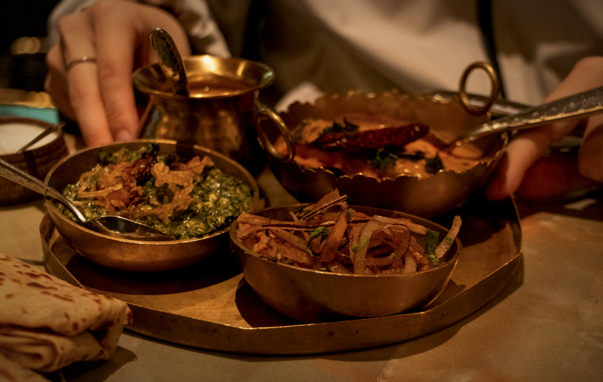 Hands serving from brass bowls of curry and dal on a brass tray, with bread at the edge of the table