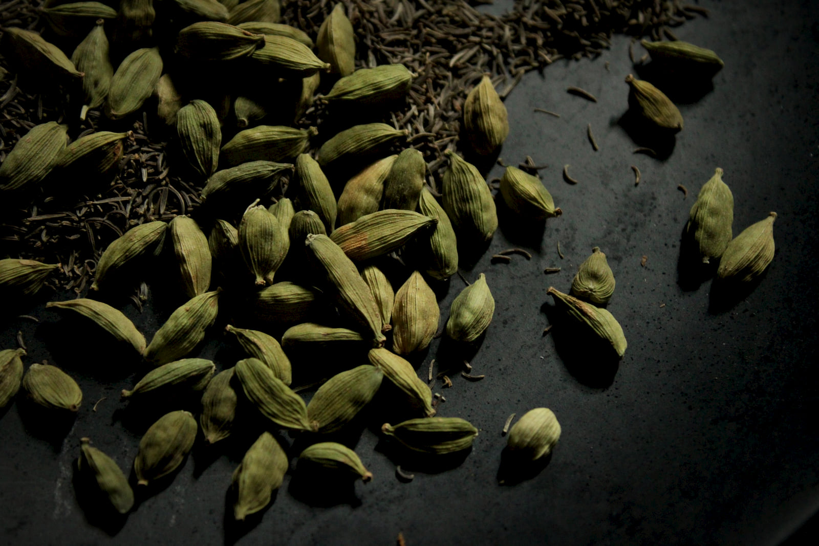 Green cardamom pods and cumin scattered on dark stone