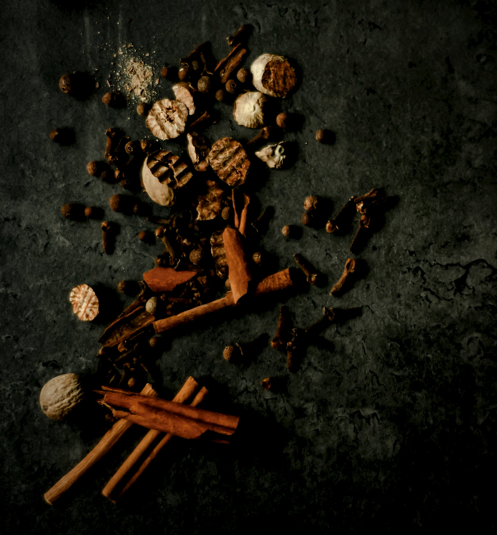Cinnamon sticks, cloves, nutmeg and mace scattered across dark stone