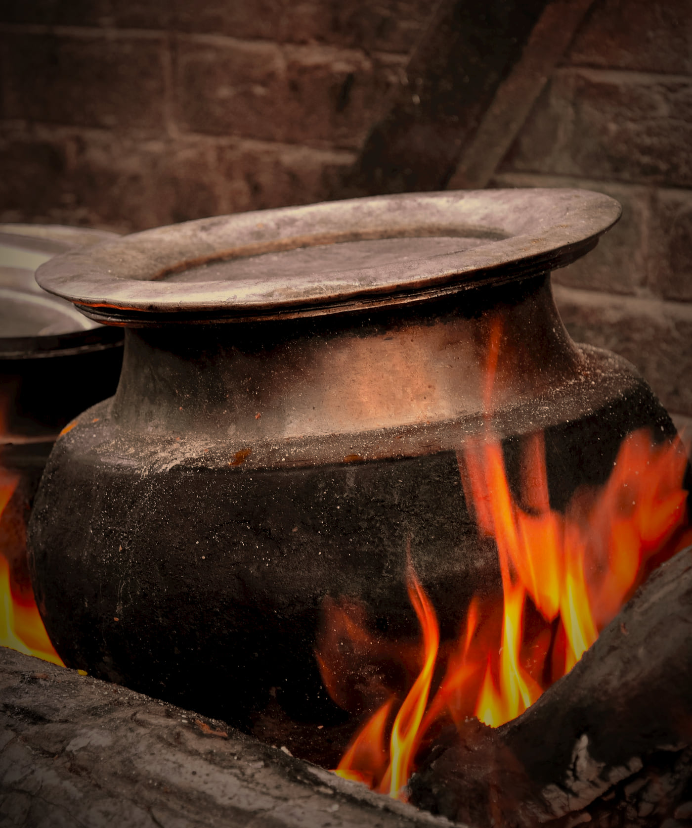 A soot-darkened handi standing in a wood fire, flames rising around its base